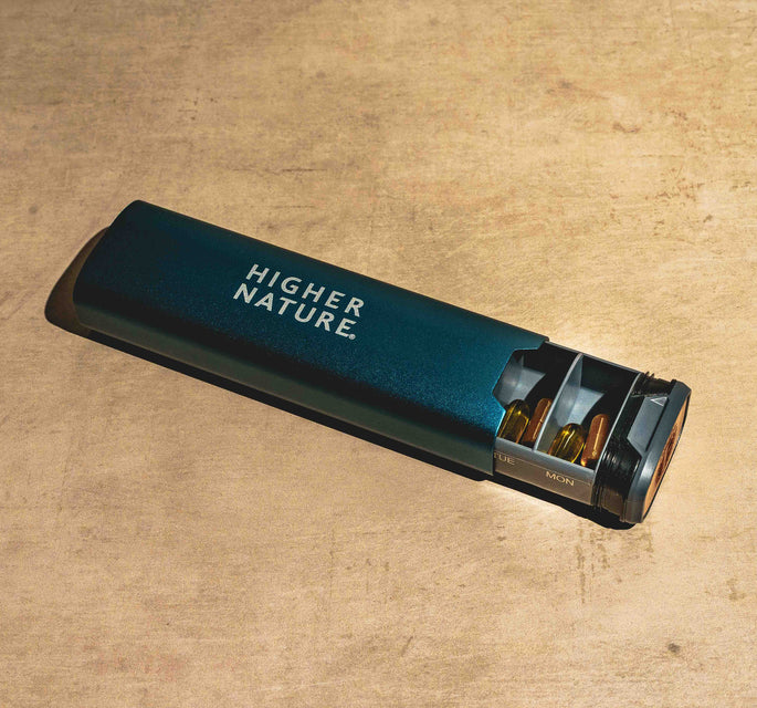 A dark teal metal lighter with the brand name 'HIGHER NATURE.' printed in white on its side, resting on a textured beige stone surface.